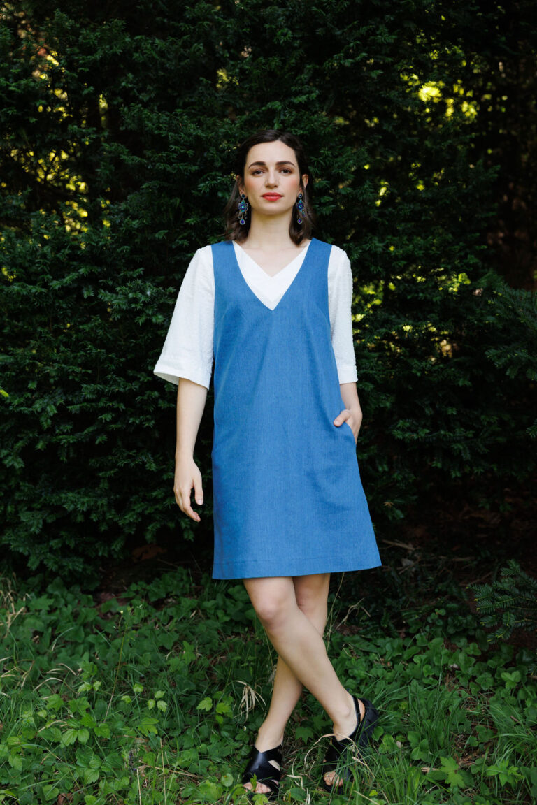 Eine Frau trägt ein schlichtes blaues Träger- Jeanskleid über einem weißen kurzärmeliger Bluse und posiert vor einem üppig grünen Hintergrund. Sie trägt auffällige blaue Ohrringe und schwarze Sandaletten, was dem Look eine stilvolle und natürliche Ausstrahlung verleiht.