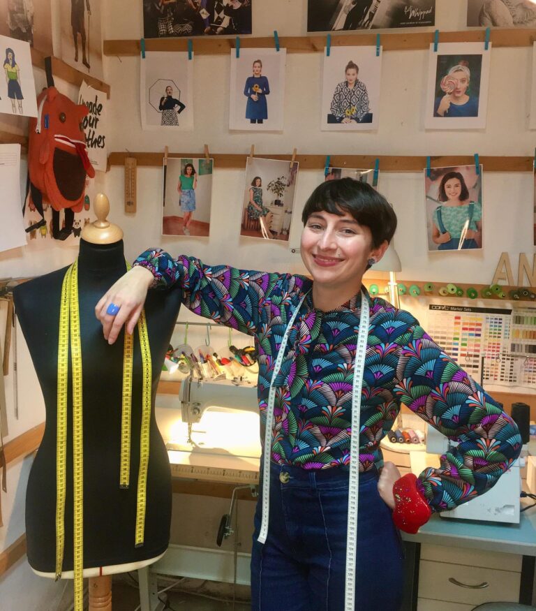 Angelika Pichler steht in ihrem Atelier mit ihrer Schneiderpuppe vor einer Nähmaschine. Man sieht viele Nähseiden und Schneiderzubehör und Fotos von vergangenden Kollektionen